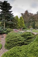 Conifers in gravel beds with ornamental sculptures beside a pond and trees beyond.  Japanese style garden, June.  North Yorkshire. 