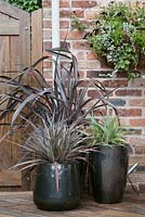 Contemporary grey and silver containers with phormium and astelia on dark stained wood decking in garden.  Brick wall and hanging basket behind.  Cheshire, June. 