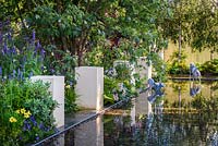 A rectangular pool with pebbles and white blocks reflected in the water in morning sunlight with wire dog sculptures in the background.  The Dog's Trust: 'It's a Dog's Life', Hampton Court Flower Show, July 2016. 