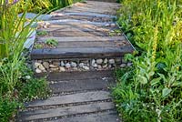 Timber and gravel path with gabion filled with stones as a step. The WWT Working Wetlands Garden. Hampton Court Flower Show 2016 - Designer: Jeni Cairn, Sponsor: Wildfowl and Wetland Trust supported by the HSBC Water Programme
