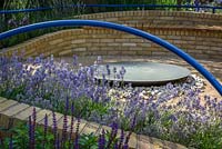Blue curving handrails above brickbenches with circular metal water feature in the centre. Plants include lavender and salvia.The Abbeyfield Society: a Breath of Fresh Air, RHS Hampton Court Palace Flower Show 2016. Design: Rae Wilkinson
