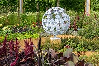 A round sculpture with leaf pattern by Paul Richardson with planting of Amaranthus 'Red Army' and purple-leaved cannas. Witan Investment Trust Global Growth Garden, RHS Hampton Court Flower Show 2016