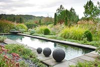 Three stoneware balls are placed by the side of the pool of the Contemporary Garden which is planted with Pennisetum alopecuroides 'Japonicum', Euphorbia cyparissias 'Fens Ruby', Cupressus sempervirens and Buxus sempervirens, in les Jardins de la Poterie Hillen.