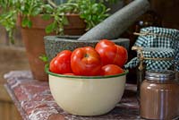 An old enamel bowl of Solanum lycopersicum, tomatoes on a stone counter top with jars of pickles and a stone mortar and pestle.
