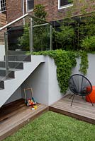 Steps with glass safety panels leading to an upper patio withchilds play area under a set of stairs with frameless glass screens a timber deck, timber children's toy and Aptenia cordifolia, heartleaf iceplant spilling over a grey painted cement rendered wall.