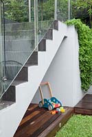 A childs play area under a set of stairs with frameless glass screens a timber deck, timber children's toy and Aptenia cordifolia, heartleaf iceplant spilling over a grey painted cement rendered wall.