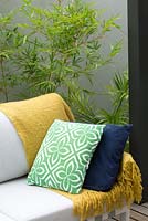 Detail of an outdoor lounge with a yellow throw rug, green and white geometric patterned and a navy blue cushion in front of a Bambusa textilis 'Gracilis', Slender weavers bamboo and a cement rendered courtyard wall.