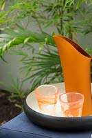 Detail of a blue outdoor table with a black and white tray an orange metal pitcher and two orange patterned drinking glasses.