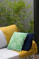 Detail of an outdoor lounge with a yellow throw rug, green and white geometric patterned and a navy blue cushion in front of a  Bambusa textilis 'Gracilis', Slender weavers bamboo and a cement rendered courtyard wall.