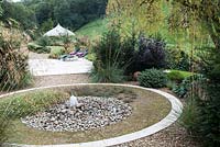 Circular water feature with water jet on pebbles and planting of Stipa gigantea Hebe and Taxus baccata