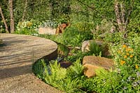 Curved path meandering among Betula nigra 'Heritage' and woodland planting and large sandstone boulders next to water rill. Vestra Wealth: Encore - A Music Lover's Garden at RHS Hampton Court Palace Flower Show 2015. Designed by Paul Martin