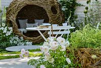 White metal bistro table and chairs and woven willow bird-hide with white and green plants including Rosa 'Iceberg - Living Landscapes 'City Twitchers' Garden, RHS Hampton Court Flower Show 2015. Designed by Sarah Keyser. CouCou Design