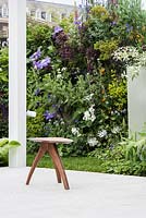 View of vertical garden next to a seating area with a wooden chair surrounded by Cotinus, Geranium 'Brookside', Alchemilla mollis, Clematis 'Patensella'. The Watahan East and West Garden.  Designers: Chihori Shibayama, Yano Tea. Sponsor: Watahan