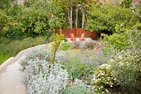 Terraced beds alongside stone steps leading to patio with bright red painted decorative chairs with Judas tree, Cistus, Stachys, Centranthus ruber, Jerusalem Sage, Papaver rupifragum and Borage