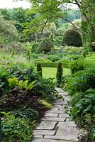 The entrance to a fifth-of-an-acre walled garden with stone path and shady border. Plants include Polygonatum x hybridum, Trachystemon orientalis, hosta, ferns and conical box. Beyond, domed topiary holly trees.