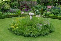 A cottage garden with circular lawn and raised wildflower mound planted with buttercups, cranesbill and cow parsley surrounding a bird bath. The formal structure is created by low box hedging and conical shaped yews.