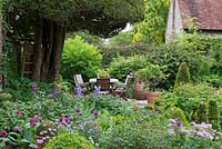 A patio with dinning table and chairs beneath a mature yew tree. In the foreground, a mixed border planted with Iris 'Jane Phillips', Allium 'Purple Sensation', roses, chives and topiary box in containers.