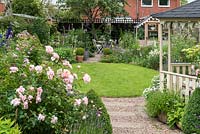 A pebble path leads past gazebo to circular lawn enclosed in cottage style herbaceous borders. At far end, there is shady seating, a compost bin amongst foxgloves, and greenhouse.