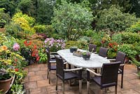 Paved, raised terrace edged in pots of dahlias, begonias, geraniums, hostas and cosmos. Dining table with potted succulents.