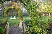 Rosa on archway over wooden bridge with decorative seat beyond in spring. Garden: Ulting Wick, Essex. Owner: Philippa Burrough