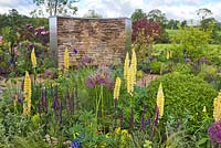 The Cruse Bereavement Care: A Time for Everything garden at RHS Chatsworth Flower Show 2017. Drystone wall with wooden bench seat, hardstanding area with island beds with Lupins, Alliums, box balls and Salvias. Design: Neil Sutcliffe