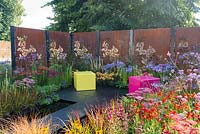 Decorative rusted steel panels enclose a colourful garden with pink and lime green cubed seating, plants include Helenium 'Moerheim Beauty', Penstemon 'Garnet', Agapanthus 'Blue Triumphator', Verbena bonariensis and Imperata cylindrica 'Rubra'- Colour Box, RHS Hampton Court Palace Flower Show 2017
