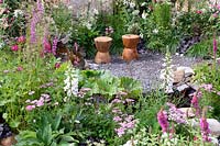 The Oregon Garden. Carved timber seats in gravel area surrounded by deep reds,cream and pink colour themed planting. Design: Sadie May Stuios. RHS Hampton Court Palace Flower Show 2017