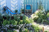 A seaside garden in front of blue and white beach huts.  Plants include: Nepeta, Double flowering Feverfew - Tanacetum parthenium 'Flore Pleno', Achillea millefolium, Verbena bonariensis and Stipa tenuissima. Designed by James Callicott Sponsored by Southend Borough Council.