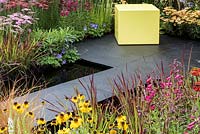 A small garden full of colourful perennials with  a pale yellow cube, two rectangular pools surrounded by slate paving. Perennials include: Achillea 'Taygatea' and 'Pink Island', Helenium 'Pipsqueak', Penstemon 'Garnet', Blood Grass - Imperata 'Red Baron' and Campanulas. Designed by Charlie Bloom.