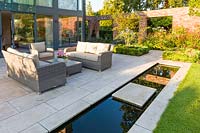 A patio area, with seating and a rill, in a modern Cheshire country garden, designed by Louise Harrison-Holland. Planting includes Amelanchier lamarckii, box hedging and Stipa tenuissima
