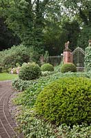A red brick path leading to classical black iron railings with stone falcons and yew topiary balls on living plinths of ivy