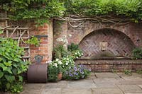 Red brick archway water feature with grouped pots of bedding plants and traditional iron lawn roller