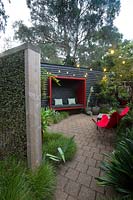 View through to paved area framed by a freestanding timber green screen planted with Muehlenbeckia complexa. A black painted timber wall with a red seating pod attached to it a red canvas retro butterfly chair and a collection of large pots with a maple and various succulents.
