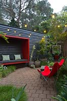 Paved area featuring a black painted timber wall with a red sitting pod attached to it a red canvas retro butterfly chair and a collection of large pots with a maple and various succulents.