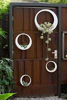 A rusty corten steel gate with a group of hand made white painted circular metal pots planted with succulents.