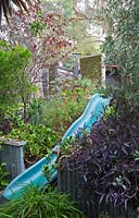 A plastic recycled slide in a sloping raised garden bed featuring Tradescantia pallida 'Purple Heart' with dark purple foliage and a mixture of succulents and shrubs.