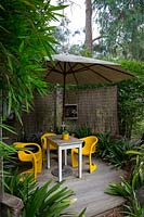 Timber deck covered by a large white umbrella with bright yellow plastic retro chairs and a timber table, screened with bamboo matchstick blinds and framed by a clump of bamboo andAspidistra elatior, cast iron plant.