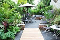 Small modern urban garden full of exotics with decking walkway over slate paving. Metal tables and chairs with a border of Tetrapanax papyrifer, syn. Aralia papyrifer, Dracaena marginata 'Tricolor Rainbow', Dicksonia antarctica, Trachycarpus fortunei, Ferns, Bamboo, Cordyline australis and Ensete ventricosum 'Maurelii'.

