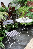 Small modern urban garden full of exotics with slate paving. Metal table and chairs with a border of bronze Phormium, Ferns, Bamboo, Cordyline australis and banana Ensete ventricosum 'Maurelii'. Pachyphytum oviferum - Moonstones - in pot on table.


