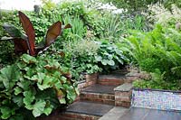 Small modern urban garden full of exotics with brick and slate steps leading to shady border.    Matteuccia struthiopteris in raised mosaic covered bed, underplanted with Saxifraga x urbium. Ensete ventricosum 'Maurelii', Ligularia dentata 'Desdemona', Phormiums, Hostas and Astilbe.