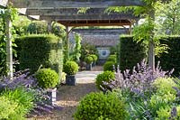 Wooden pergola over formal pathway with clipped topiary in containers, Nepeta 'Six Hills Giant' and Alchemilla mollis