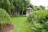 A woven Willow structure with Birch trees and summerhouse in background