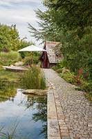 A granite paved path edging the natural swimming pond goes to the witches' cottage situated next to a wooden deck with umbrella. Cedrus libani