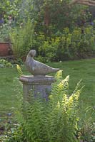 Decorative bird stone statue amongst ferns in spring. Garden: Quarry Cottages, Sussex