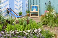 Blue and white painted beach huts and deck chairs on decking, driftwood posts, gravel, pebbles and planting of grasses, Eucalyptus gunnii 'Azura' underplanted with Achillea ptarmica 'The Pearl', Achillea millefolium 'Wonderful Wampee', Nepeta racemosa 'Walker's Low', Verbena bonariensis, Stipa tenuissima in front of blue fence - By The Sea - RHS Hampton Court Palace Flower Show 2017 - Design: James Callicott -Sponsor: Southend Borough Council.