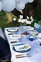 Outdoor dining table dressed in shades of blue and a vase of white Japanese anemones.  Paper ball lanterns and paper pompoms hang over the table to decorate. Pockets are made in the folded napkins to put name card and each setting is finished with an olive branch