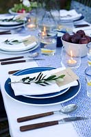Outdoor dining table dressed in shades of blue and with a vase of Japanese anemones. Pockets are made in the folded napkins to put name card in and each setting is finished with an olive branch. Tealights in glasses light the table as dusk falls