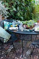 Outdoor dining area under a fig tree on a brick patio. Table laid with greens and fresh whites and napkins dressed with a geranium leaf