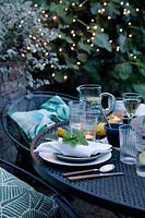 Dusk falls on the outdoor dining area under a fig tree. Fairy lights are in the trees creating a cosy setting and light up the area. Tealights have been lit on the table which is laid with greens and fresh whites and napkins dressed with a geranium leaf