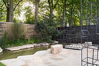 View of patio and water feature and metal sculptures surrounded by Betula pendula, Pinus sylvestris - Breaking Ground - RHS Chelsea Flower Show 2017 - Designers: Andrew Wilson and Gavin McWilliam - Sponsor: Darwin Property Investment Management Ltd